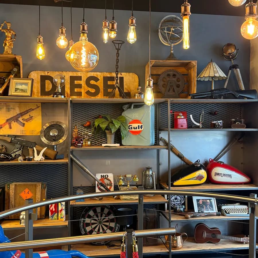 Regal unter warmen Glühbirnen mit Vintage-Deko: Holzschild „Diesel“, Benzinkanister, zwei alte Motorrad-Tanks und eine Dartscheibe