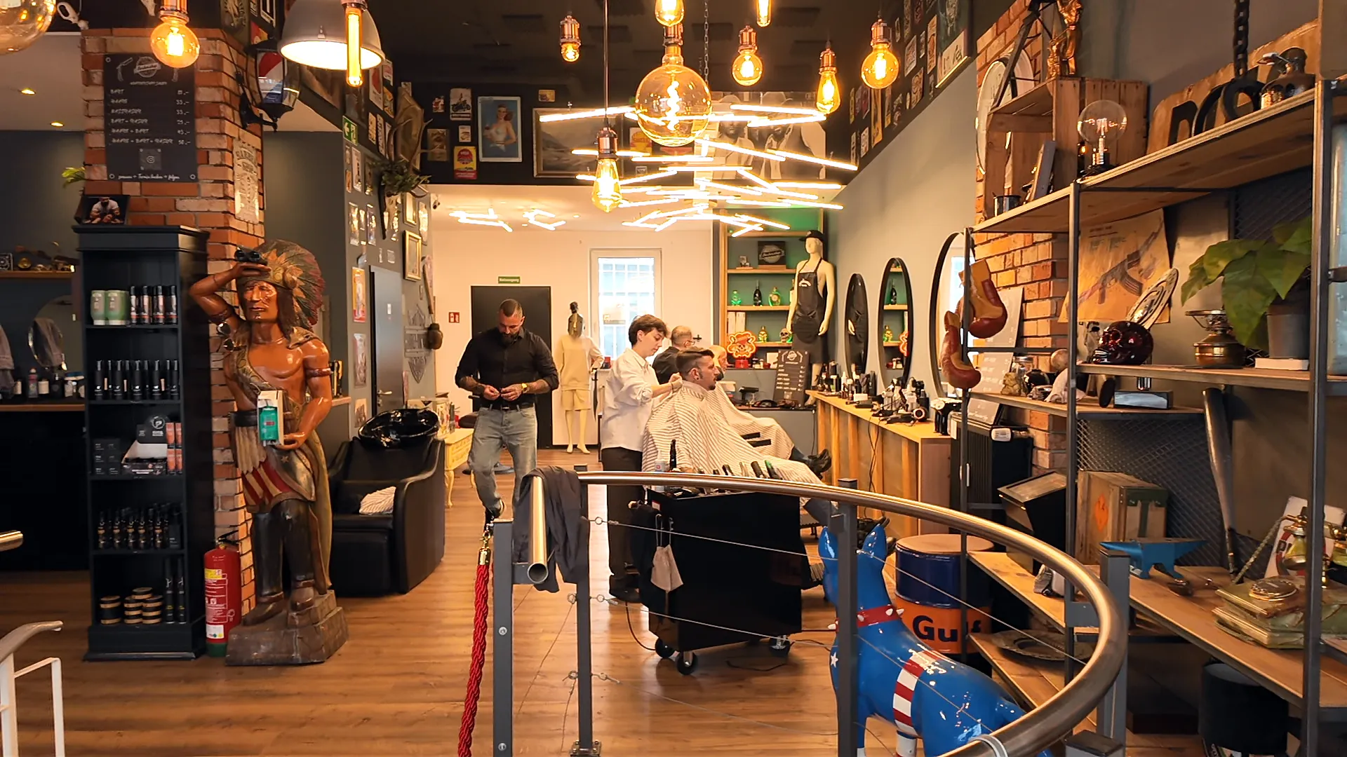 Blick durch den Barbershop Sauer in Bielefeld: geschnitzte Holzfigur, Regale, warme Glühbirnen, hinten Barber bei der Arbeit