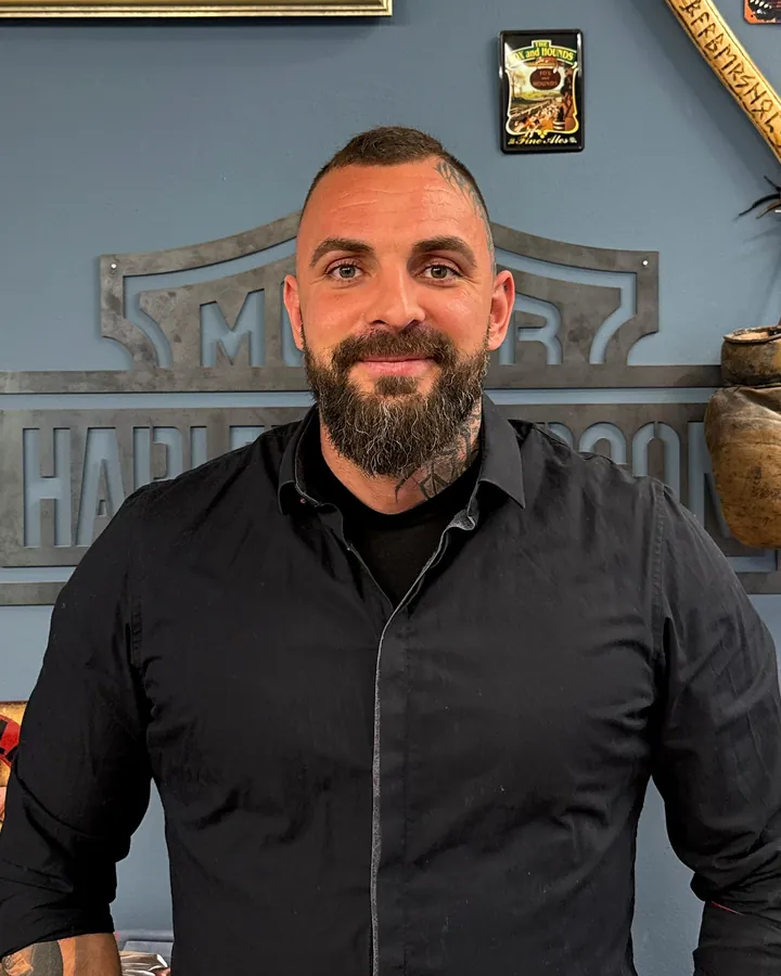 Richard, Inhaber vom Barbershop Sauer, mit Vollbart und schwarzem Hemd vor dem Harley-Davidson-Schild im Laden